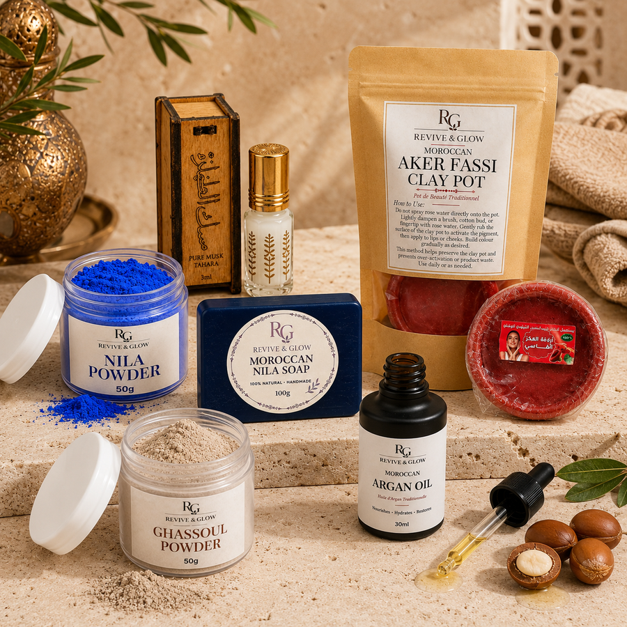 A curated product arrangement on a beige stone surface featuring Revive Reglon skincare items: a clay pot, powder jars, soap bar, argan oil bottle, and wooden box, with a red bath bomb dish and small green leaves as styling props.