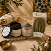 A styled spa product arrangement on a stone slab featuring a jar of black balm soap, a jar of terracotta powder, and a packaged exfoliating glove by Kiss Medical, with folded textiles and decorative brass vessels in the background.