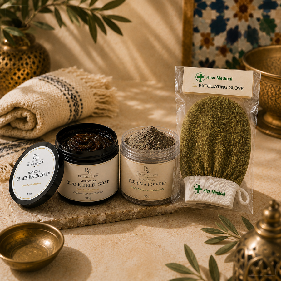 A styled spa product arrangement on a stone slab featuring a jar of black balm soap, a jar of terracotta powder, and a packaged exfoliating glove by Kiss Medical, with folded textiles and decorative brass vessels in the background.