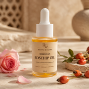 A 30ml bottle of Revive & Glow Moroccan Rosehip Oil with a white dropper cap, styled on a beige fabric surface with a pink rose, rose petals, and rose hips.