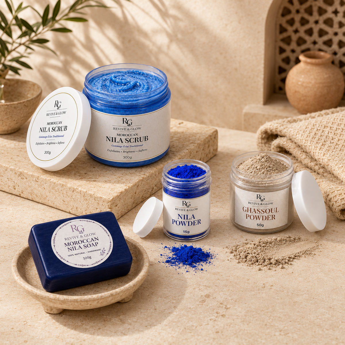 A styled product display of Revive & Glow Skin Moroccan Nila Scrub and Nila Powder. The set includes a deep blue bar of soap in a wooden dish, a blue scrub in a glass jar with its lid off, a jar of blue powder, a jar of beige glassoul powder, and a white-lidded jar of Moroccan Nila Scrub, all arranged on a beige stone surface with a burlap cloth and decorative pottery in the background.