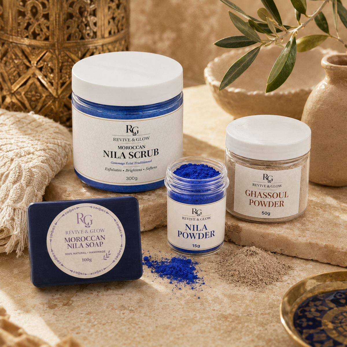 A styled product arrangement of Revive & Glow Moroccan Nila Scrub, Moroccan Nila Soap bar, and Moroccan Nila Powder in jars and a small jar, set on a textured beige surface with stone, linen, and gold decorative elements.