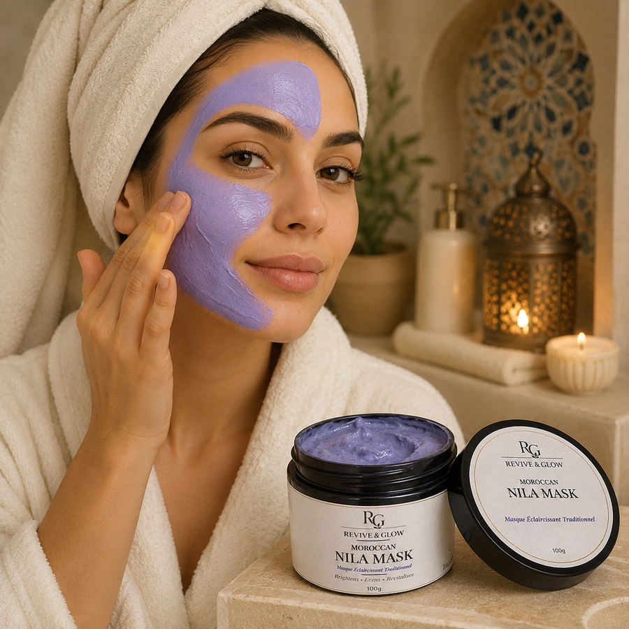A woman in a white bathrobe with a white towel wrapped around her head applies a lavender-purple face mask to her cheek. In the foreground, a jar of Revive and Glow Moroccan Nila Mask is shown with its lid beside it, set against a warm, spa-like background with candles and decorative lanterns.