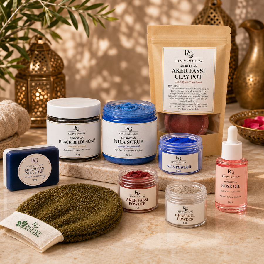 A curated spa gift set from Revive & Glow arranged on a marble surface with warm decorative lighting. The set includes a beige clay pot, several labeled jars of soap, scrub, powder, rose oil, a green towel, a blue soap bar, and a small pouch, all presented together as a coordinated product offering.